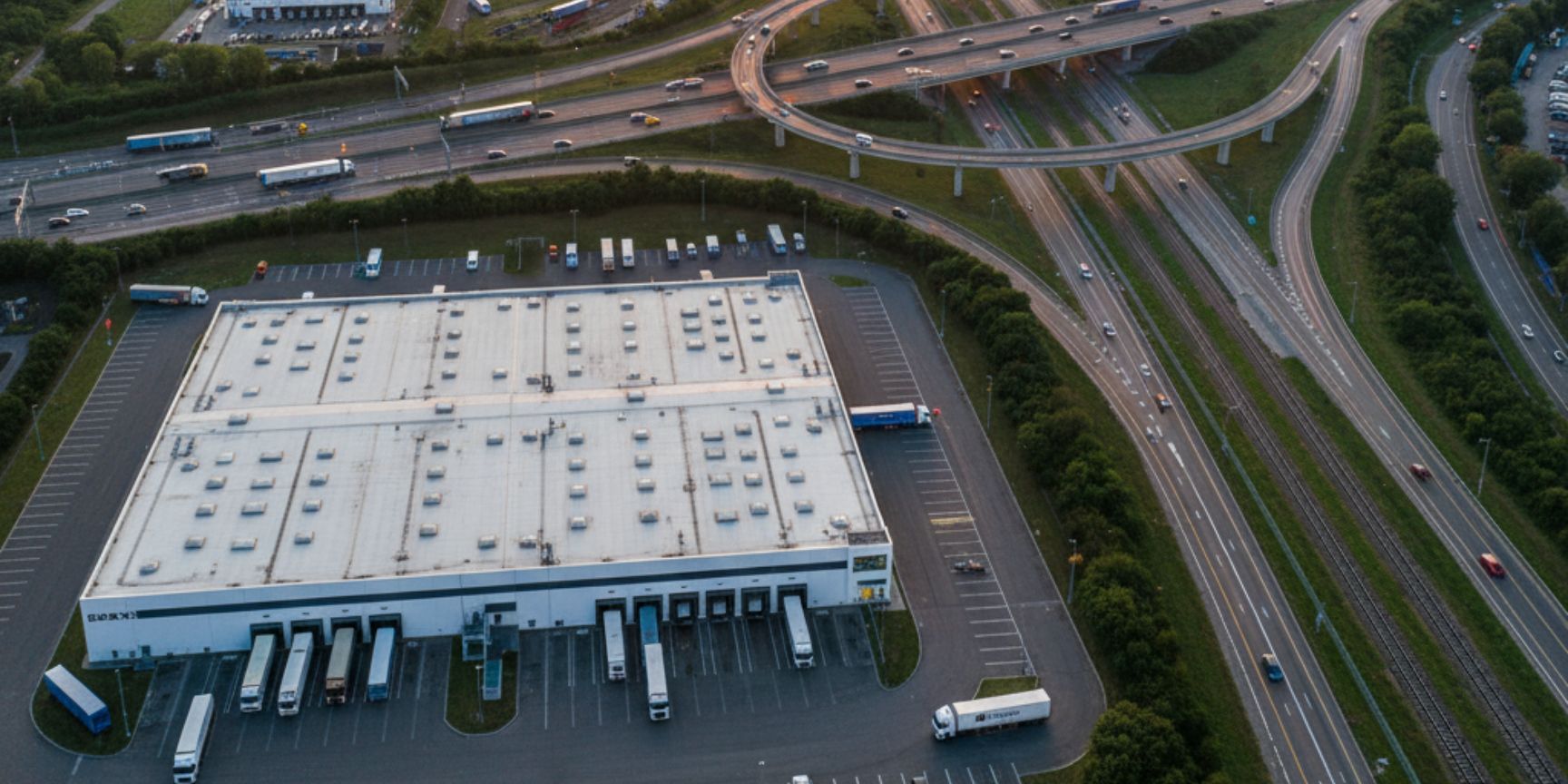 Drone shot of a large logistics center at sunset, surrounded by highway interchanges with flowing traffic, freight railway tracks and a container port in the background. The scene shows the networking of transport routes and logistics properties in an urban environment, symbolizing the meso-logistics level